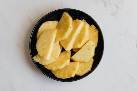 Top view of black ceramic bowl with slices of fresh sweet yellow pineapple placed on marble surface